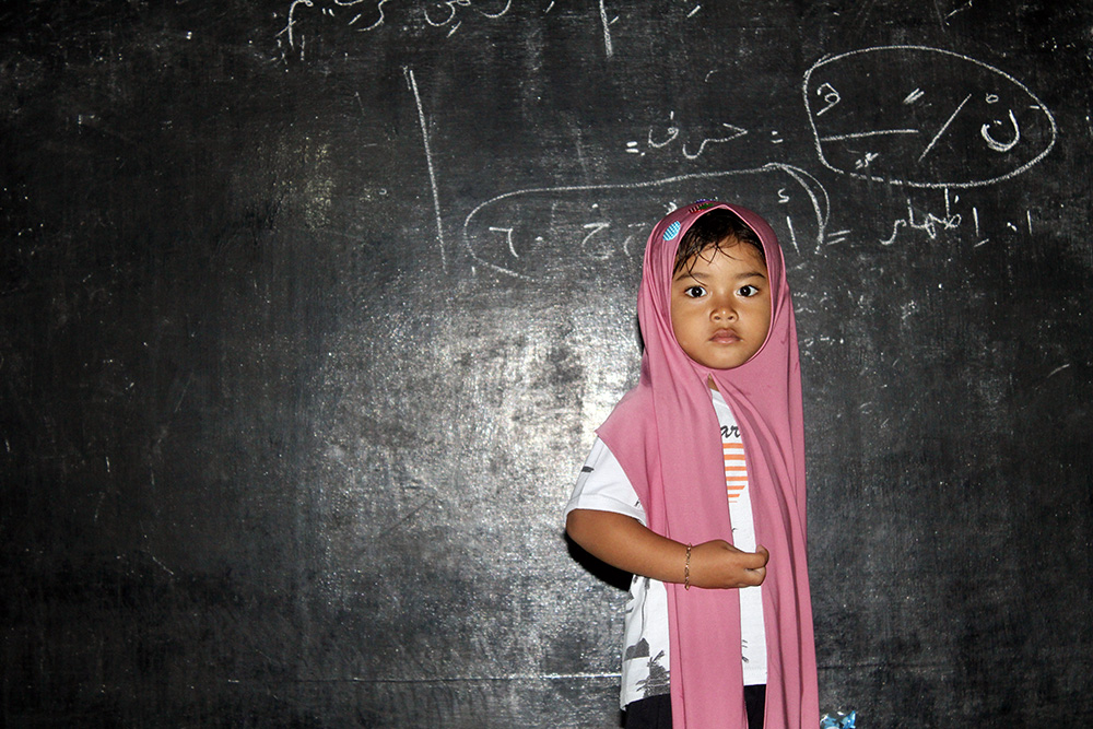 A young child wearing a headscarf standing in front of a blackboard